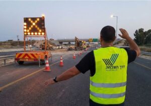 פקח תנועה של חברת דרך הצלחה מכוון את התנועה באתר עבודות תשתית בכביש מהיר בשעות השקיעה עם עגלת חץ מוארת.