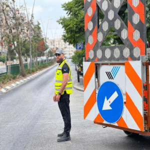 פקח תנועה מוסמך של חברת דרך הצלחה עומד ליד עגלת חסימה ממותגת עם תמרור חץ שמאלה, המכוון את התנועה בכביש עירוני.